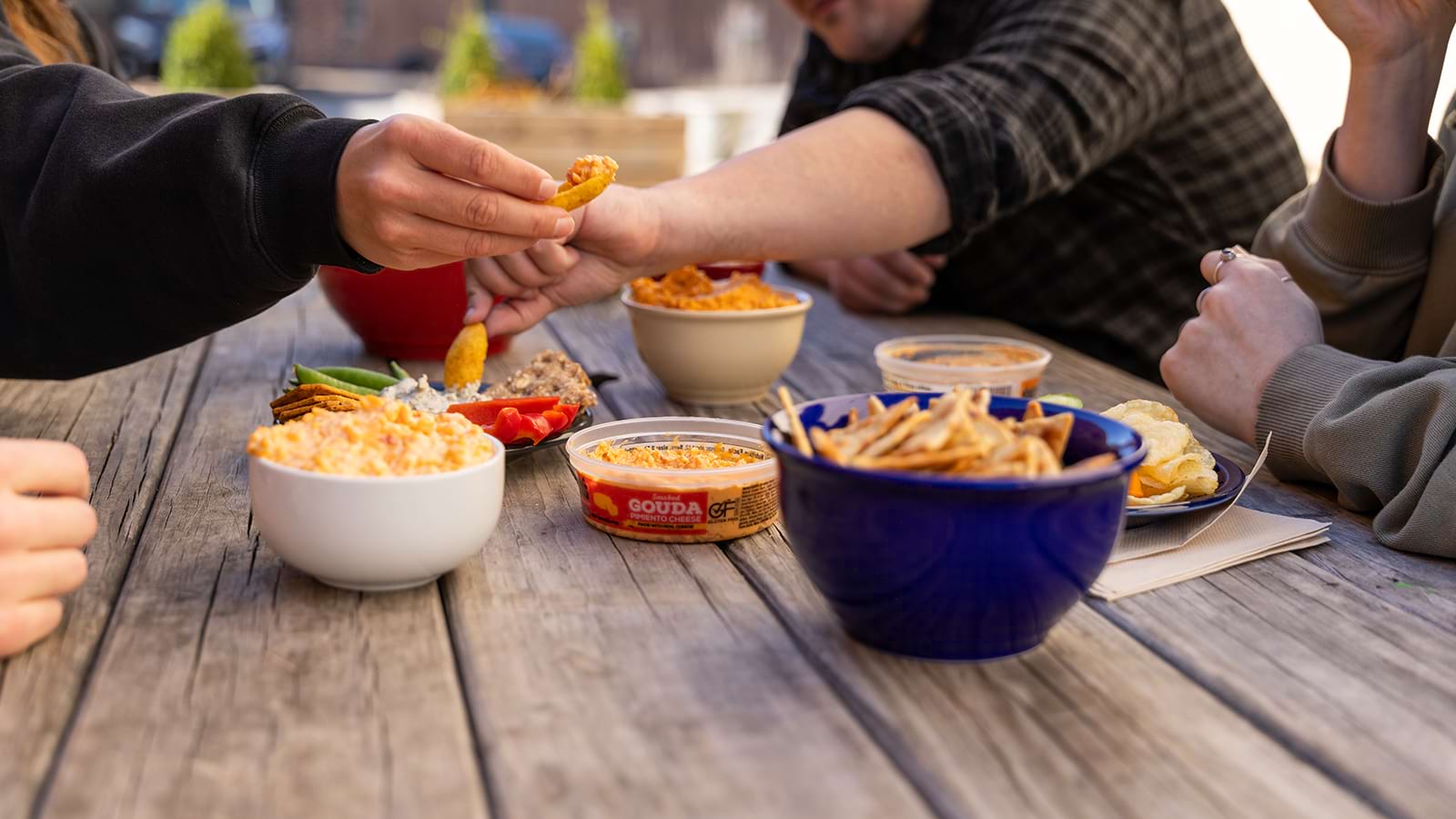 individuals gathered around a wooden table and reaching into Knott's dips and spreads with a variety of chips, veggies and crackers