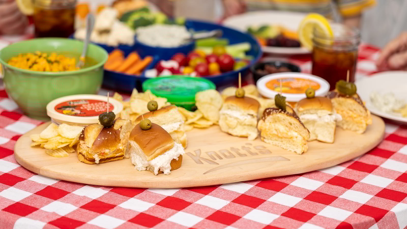 Knott's sliders on a tray with Knott's bacon cheddar ranch dip, jalapeno popper dip and spicy chicken taco dip, with veggies in the background
