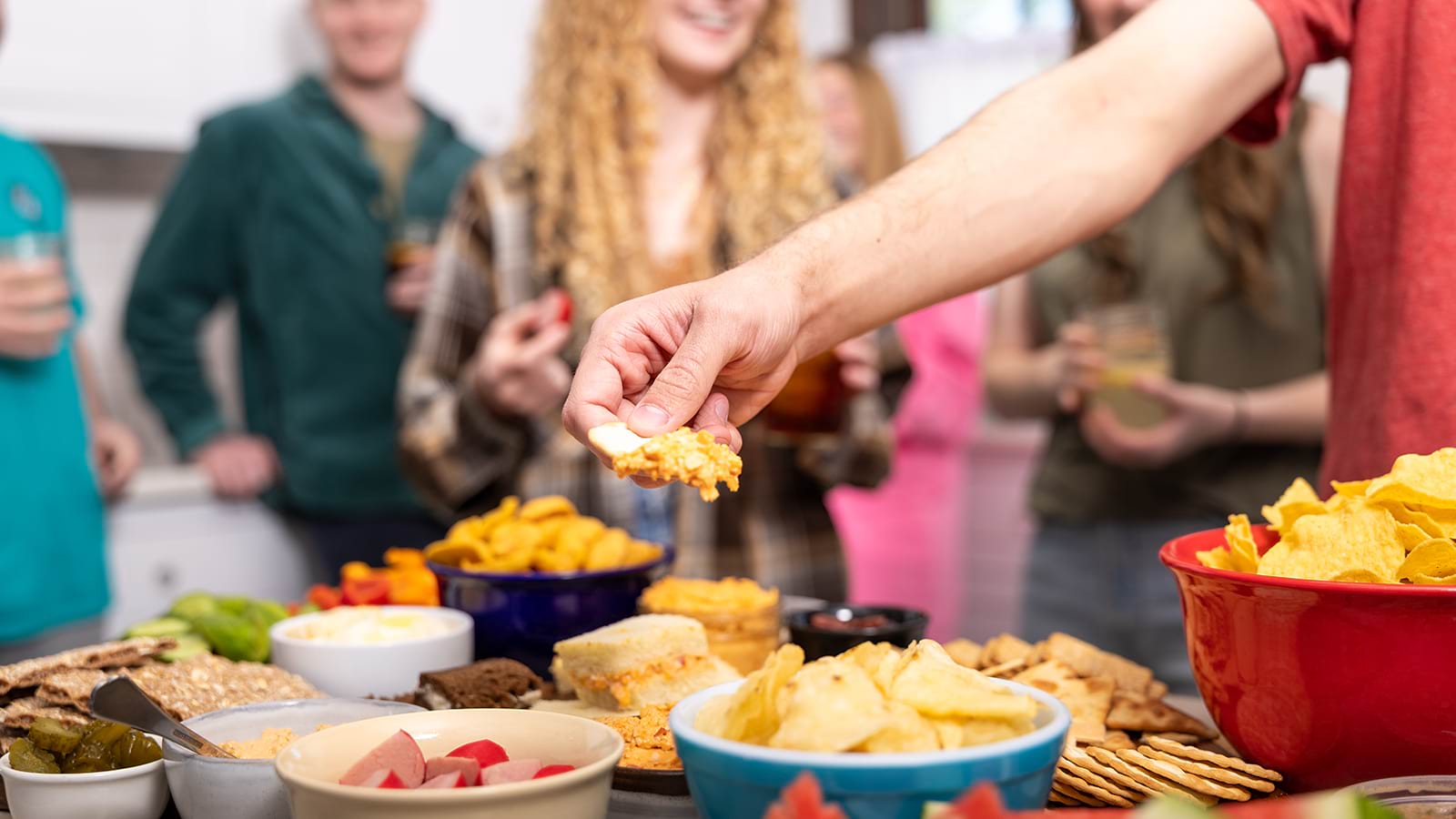 A mans arm and hand dipping into knott's chicken buffalo dip while his friends are in the background talking and smiling