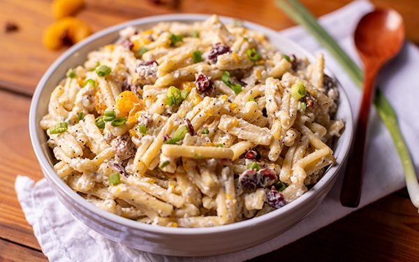 autumn chicken pasta salad with poppy seeds, cranberries and oranges in a bowl with a wooden spoon and green onion stem on the side