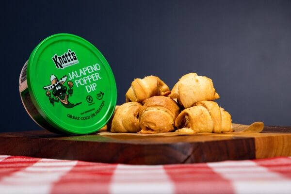 jalapeno popper pigs in a blanket stacked on a wooden board next to a container of knotss jalapeno popper dip